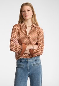 Blusa marrone a fantasia con chiusura a bottoni e maniche oversize, abbinata a pantaloni in denim azzurro chiaro. Texture liscia.