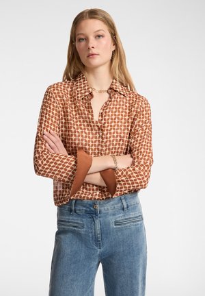 Blusa marrone a fantasia con chiusura a bottoni e maniche oversize, abbinata a pantaloni in denim azzurro chiaro. Texture liscia.