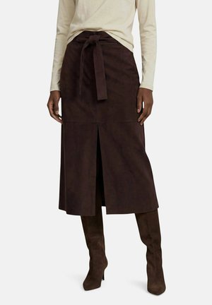 Femme portant une jupe midi marron foncé avec une fente devant et une ceinture nouée à la taille, associée à des bottes hautes assorties et un haut léger à manches longues.