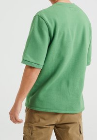 Zelené tričko s krátkým rukávem a kulatým výstřihem, vyrobené z bavlny. Obsahuje ohrnuté manžety a detailem na zadním díle. Stylizováno s khaki cargo kalhotami.