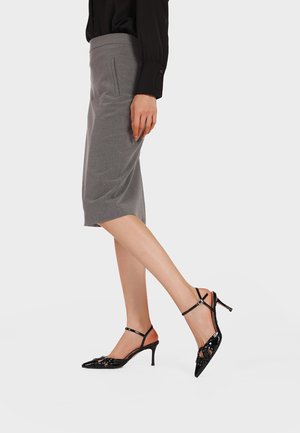 Donna con gonna grigia al ginocchio e camicetta nera, indossa décolleté slingback nere in vernice con punta a punta e sottili cinturini.