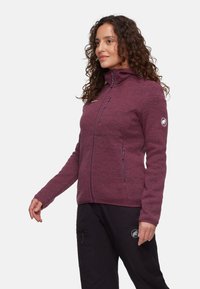 Bordowy polarowy kurtka z wysokim kołnierzem, zapinanymi kieszeniami i naszywką z logo na rękawie. Posiada teksturowany materiał i dopasowany krój.