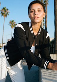 Mladić se naslanja na betonsku ogradu na otvorenom, nosi crnu koledžicu s bijelim prugama i bijele hlače, u pozadini su palme.