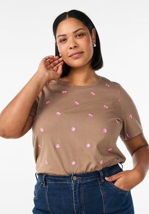 Kobieta o czarnych włosach do ramion ubrana w brązową koszulkę z małymi różowymi kwiatami i niebieskie dżinsy, pozująca z jedną ręką przy uchu.