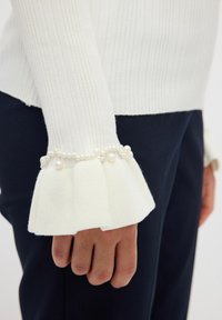 Weißer gerippter Pulloverärmel mit weitem Bund, der mit Perlendetails versehen ist. Der Bund hat ein strukturiertes Design, das das visuelle Interesse erhöht.