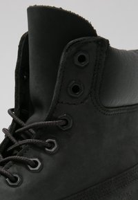 Gros plan sur la partie supérieure d'une botte en cuir noir montrant les œillets, les lacets et le col rembourré sur un fond uni clair.