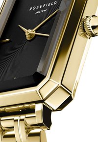 Reloj rectangular dorado con una esfera negra texturizada y líneas concéntricas, manecillas doradas y una caja pulida. Detalle notable de la corona visible.