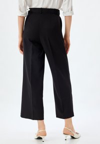 Pantalons noirs taille haute avec une coupe droite, dotés d'une ceinture, de poches latérales et d'une texture lisse. Des chaussures à talons hauts blanches complètent le look.
