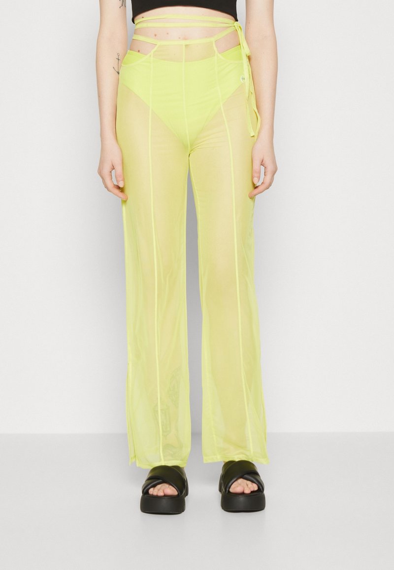 Femme portant un pantalon jaune néon transparent taille haute avec une ceinture à nouer et des sandales à plateformes noires, debout sur un fond blanc uni.