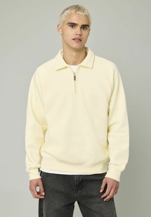 Jeune homme aux cheveux courts décolorés portant un sweat-shirt léger jaune pâle à col zippé et un jean gris foncé, le tout sur un fond gris uni.