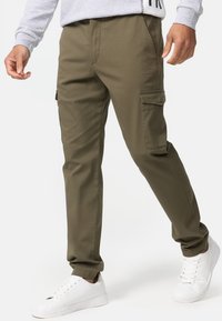 Pantalones cargo verde oliva con bolsillos laterales, fabricados en una mezcla de algodón. Presentan un fit entallado y una textura suave. Llevados con zapatillas blancas.