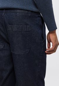 Tmavomodré rifľové nohavice s bočnými a zadnými vreckami, s jemnou textúrou. Pás má pútka na opasok a čistý zakončenie.