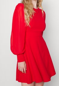 Punainen mekko, jossa on puffattuja läpikuultavia hihoja, tiivis vartalo ja leveä hame. Sileä, yksinkertainen kangas. Minimalistinen muotoilu ilman koristuksia.