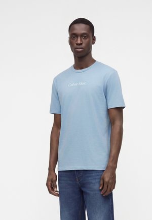 Jeune homme portant un t-shirt Calvin Klein bleu clair et un jean bleu, debout devant un fond blanc uni.