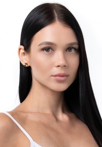 Goldene, herzförmige Ohrringe mit strukturiertem Finish, präsentiert vor einem neutralen Hintergrund. Das Modell hat langes, glattes schwarzes Haar.