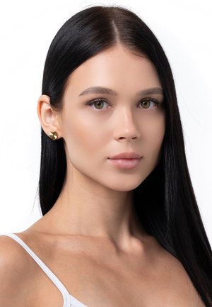 Goldene, herzförmige Ohrringe mit strukturiertem Finish, präsentiert vor einem neutralen Hintergrund. Das Modell hat langes, glattes schwarzes Haar.