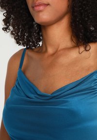 Turquoise blus med hög krage och tunna axelband, gjord av mjukt material; har en draperad design och en slät yta.