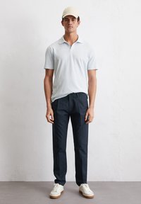Lichtblauw polo shirt met een kraag, marineblauwe broek en witte sneakers. Buitenscène met een gearticuleerde lichte achtergrond.
