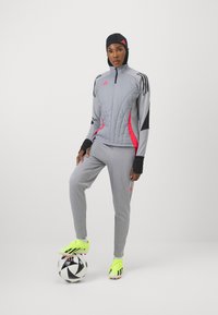 Chándal deportivo gris con acentos rosas; parte superior acolchada, ribete negro, mangas largas, guantes. Botas de fútbol amarillas brillantes, de pie sobre un balón en blanco y negro.