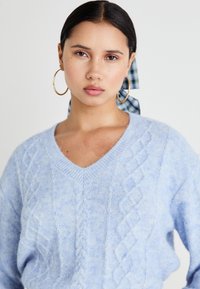 Hellblauer Kabelstrickpullover mit V-Ausschnitt und weicher Textur. Das Model trägt goldene Creolen und ein kariertes Band im Haar.
