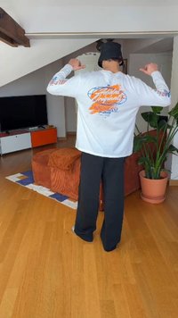 Hosszú ujjú fehér ing színes grafikával a háton, fekete nadrággal párosítva. Szoba fa padlóval, növénnyel és egy televízióval.