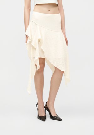 Frau trägt einen asymmetrischen cremefarbenen Rock mit gestuften Rüschen, kombiniert mit schwarzen spitzen High Heels, vor einem schlichten Hintergrund.