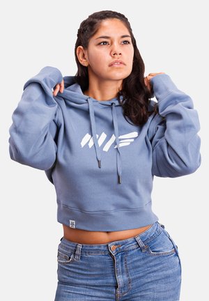 Blue cropped hoodie with drawstrings, elastic waistband, and white logo. Soft material with long sleeves; paired with high-waisted jeans.
