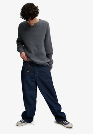 Person iført en grå strikket sweater, mørke løstsiddende jeans med nøgler fastgjort og mørke sneakers, kigger ned med hænderne i lommerne.