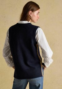 Granatowy dziergany kamizelka z prążkowanym ściągaczem i brązowymi akcentami po bokach, zestawiona z jasnym koszulą z marszczonymi rękawami.