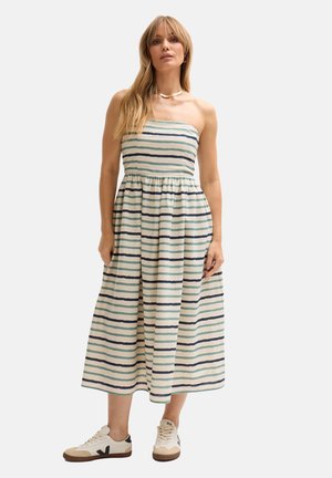 Frau trägt ein trägerloses, gestreiftes Midikleid und weiße Turnschuhe, steht mit entspannten Armen an den Seiten vor einem schlichten Hintergrund.