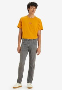 T-shirt en coton jaune avec un logo Levi's rouge, associé à un jean en denim gris et des baskets blanches. Le modèle se tient devant un fond uni.