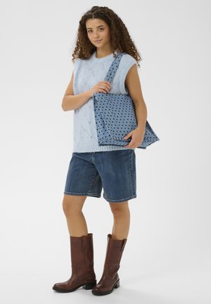 Junge Frau trägt einen hellblauen ärmellosen Pullover, Jeansshorts, braune Stiefel und hält eine blaue Tragetasche mit schwarzen Herzmustern.