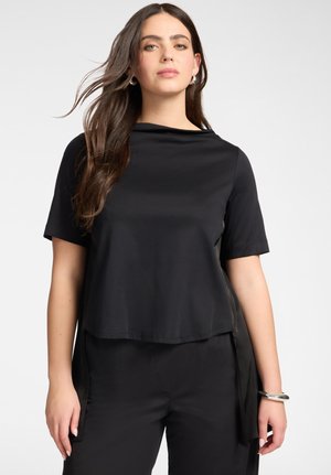 Młoda kobieta z długimi brązowymi włosami nosi czarny krótki top i spodnie, stoi na prostym jasnym tle.