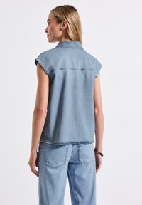 Top in denim senza maniche di colore azzurro, con collo a camicia, caratterizzato da una linea dritta e dettagli di cucitura minimi sul retro. Abbinato a jeans azzurri.