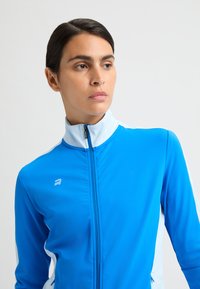 Chaqueta deportiva azul con cremallera y cuello alto, hecha de una tela suave y elástica. Presenta un acento blanco en las mangas y un logo en el pecho.