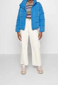 Blå puffjacka med dragkedja och huva, parat med en randig topp, vita vida byxor och beige sneakers. Mjuk tyg, avslappnad stil.