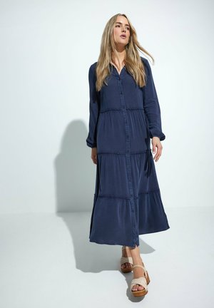 Robe maxi en tissu bleu marine foncé, avec un devant à boutons, des manches longues et un design à volants. Portée avec des sandales à plateforme beige clair.