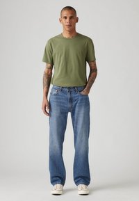 Νεαρός άνδρας με τατουάζ, φορώντας ελαιόχρωμο μπλουζάκι, μπλε τζιν και λευκά αθλητικά παπούτσια, στέκεται μπροστά από απλό ανοιχτό γκρι φόντο.