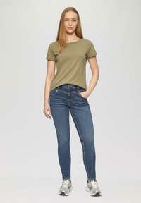 T-shirt ample de couleur vert olive, ajusté. Jeans skinny bleu foncé avec poches avant et détails de couture. Chaussures de sport argentées.
