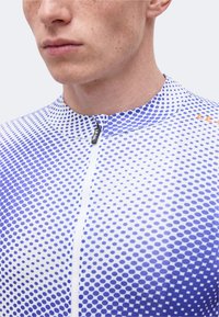 Junger Mann, der ein weiß-blau gepunktetes Radtrikot mit Frontreißverschluss und einem kleinen Stoffetikett am Kragen trägt.