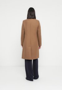 Femme avec des cheveux noirs raides portant un long manteau en camel, un pantalon large noir, se tenant contre un fond clair avec le dos tourné vers le spectateur.