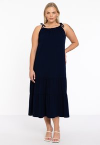 Navyblaues Maxikleid aus weichem Stoff mit verstellbaren Bindeträgern, einem gestuften Rüschendesign und einer lockeren Passform. Sandalen mit flacher Sohle.