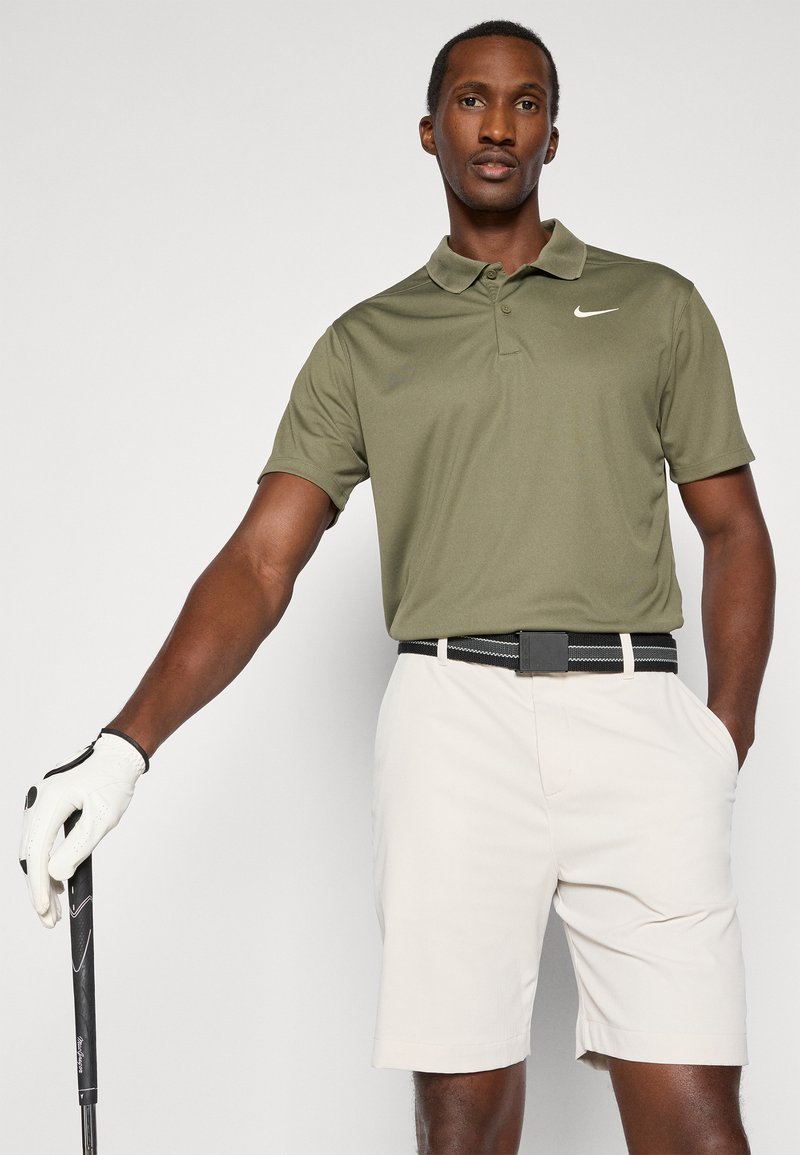 Polo shirt Nike pour hommes en vert olive, avec un col et des manches courtes. Associé à un short beige et une ceinture noire. Club de golf à la main.