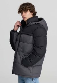 Giovane uomo che sistema il colletto di un giubbotto imbottito bicolore nero e grigio con cappuccio, indossando jeans azzurri, contro uno sfondo chiaro e semplice.