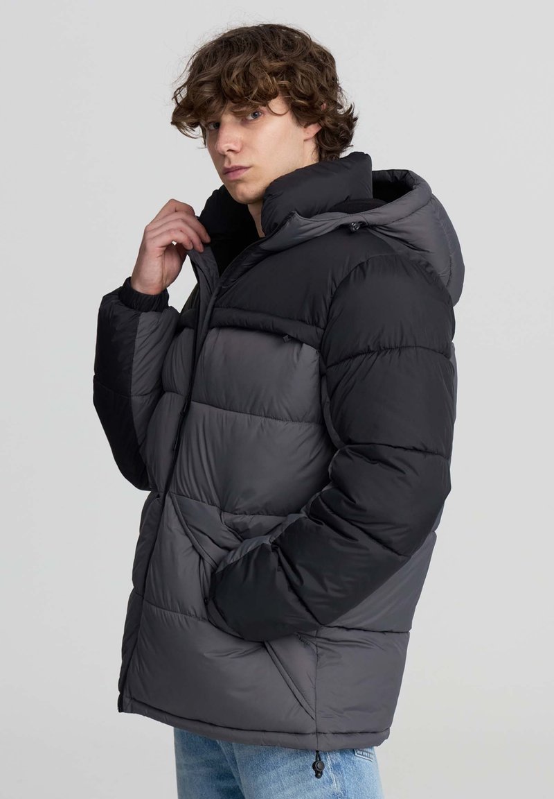 Giovane uomo che sistema il colletto di un giubbotto imbottito bicolore nero e grigio con cappuccio, indossando jeans azzurri, contro uno sfondo chiaro e semplice.