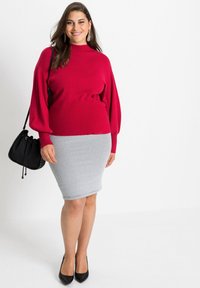Maglione rosso lavorato a maglia con collo alto, abbinato a una gonna a tubino grigia con texture. Una borsa nera e décolleté con punta completano l'outfit.