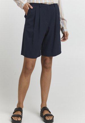 Shorts plissés bleu marine en tissu léger, avec une coupe ajustée et des poches latérales, associés à des sandales noires.