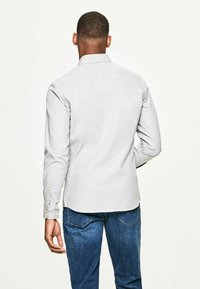 Camicia a maniche lunghe grigia chiara con polsini abbottonati e colletto classico. Il retro ha una texture liscia e un orlo curvo. Abbinata a jeans blu.