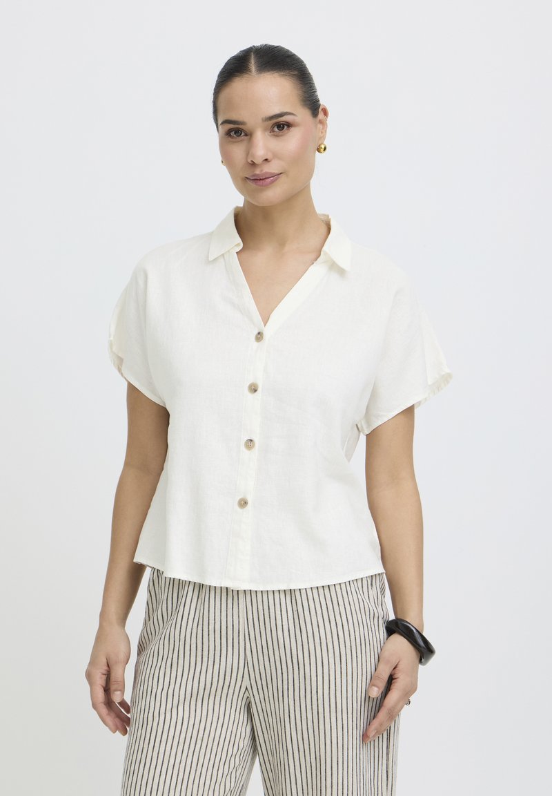 Femme portant une chemise blanche à manches courtes boutonnée, un pantalon rayé, des boucles d'oreilles en or et un bracelet noir, debout devant un fond uni.
