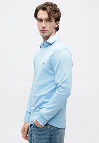 Fiatal fiú, hullámos barna hajjal, világos kék pulóverben, ing fölött és kék farmerben, áll egy sima, fehér háttér előtt.